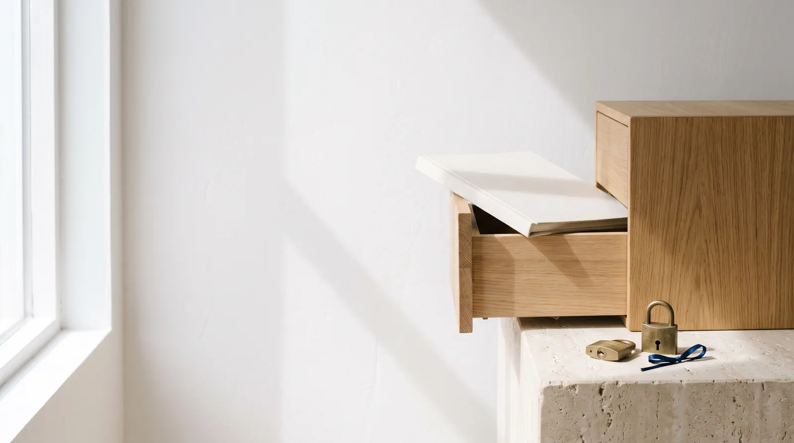 A light oak drawer half open with a white document folder, a brass padlock and a small navy ribbon on a travertine block.